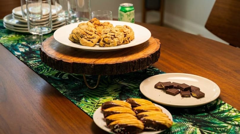 A wooden table with a tropical runner, displaying cookies, chocolates, and glasses.