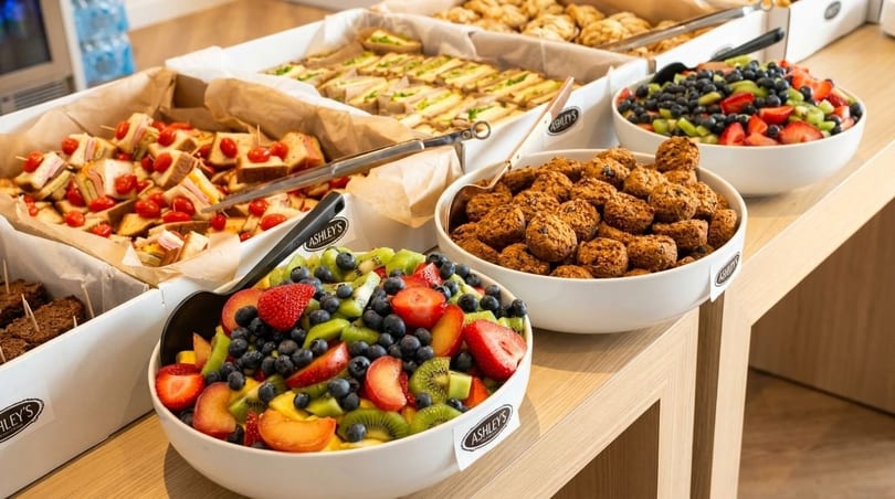 A buffet spread with sandwiches, fruit salad, and cookies in white bowls on a wooden table.