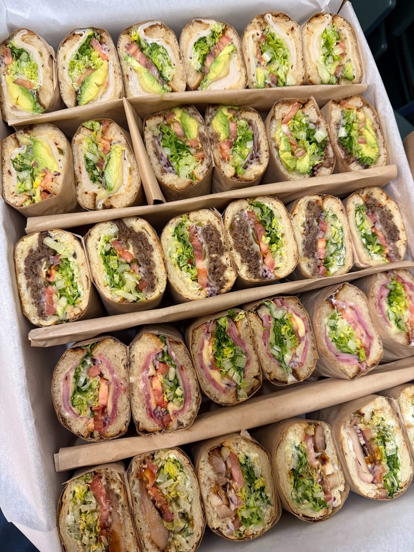 A catering box filled with rows of various submarine sandwiches, each cut in half and wrapped.