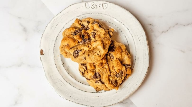 Two golden-brown chocolate chip cookies with melted chips on a rustic white plate.